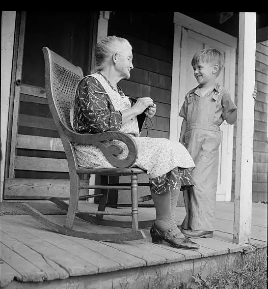 older woman sitting in rocking chair and knitting talking to a young boy on the porch